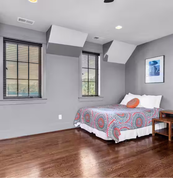 The Glass House bedroom with hardwood floor, gray walls, two windows with blinds, a bed with a colorful patterned quilt and orange decorative pillow, and a wooden nightstand.
