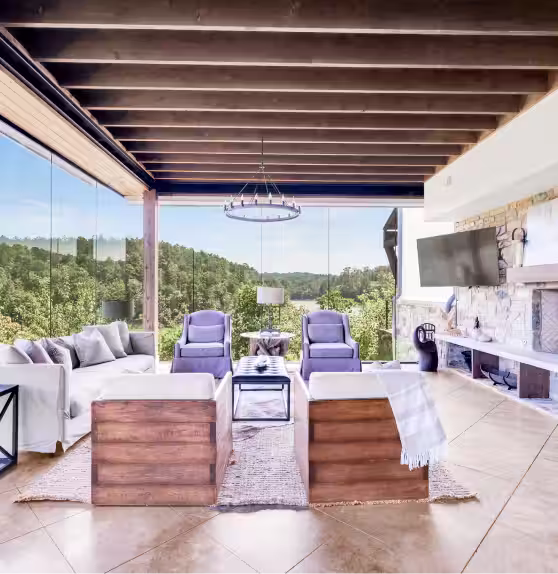 The Glass House spacious covered patio with wooden ceiling beams, two wooden armchairs, two purple chairs, a gray sofa, and a mounted flat-screen TV overlooking a lush green lake view.
