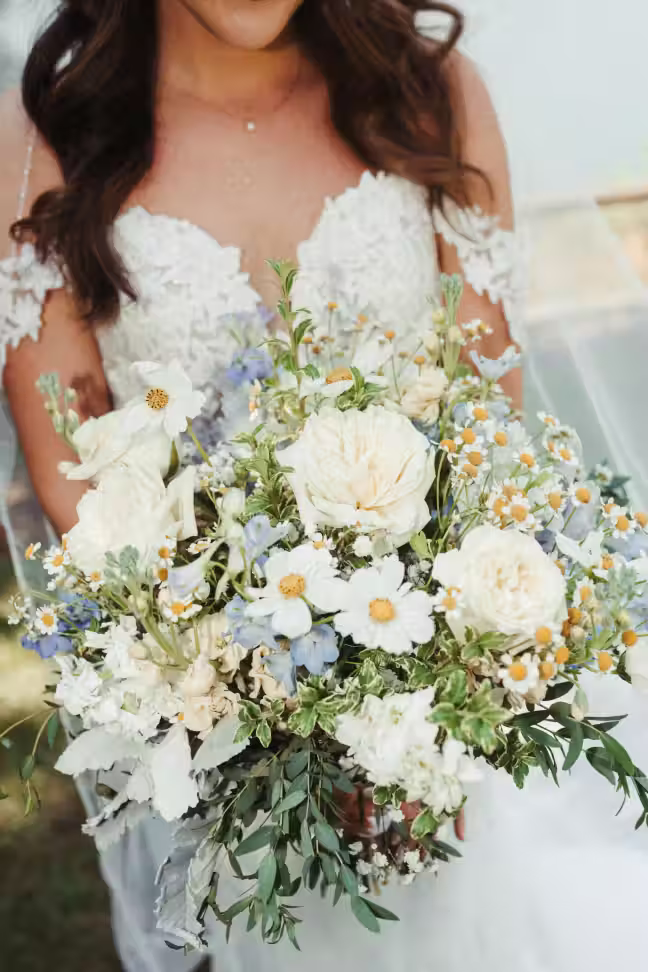 Bride in a white lace wedding dress holding a bouquet of white and light blue flowers with greenery.
