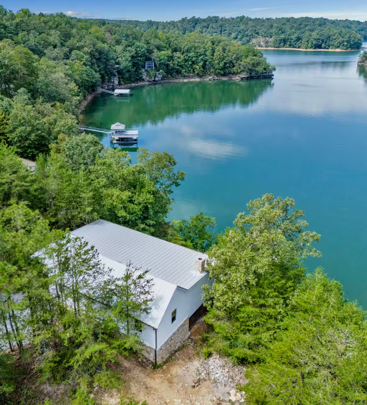 White cabin with a metal roof surrounded by green trees beside a calm lake with forested hills in the background.