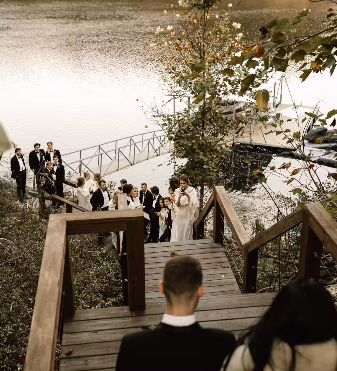 Guests in formal attire gathered by a lake dock surrounded by autumn foliage, viewed from a wooden staircase.