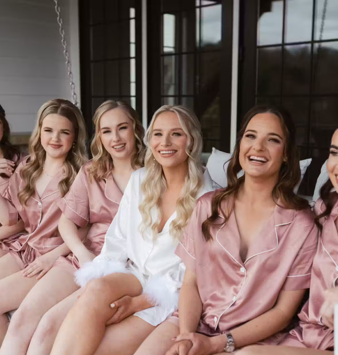 Group of smiling women sitting side by side wearing matching pink satin pajamas, with one woman in white satin robe with feathery cuffs.