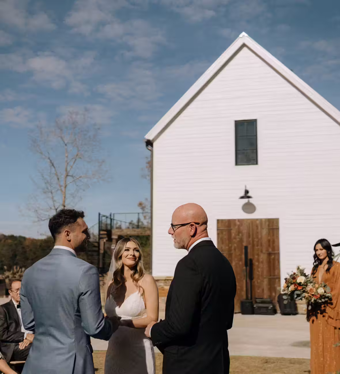 Bride and groom holding hands during outdoor wedding ceremony with officiant and bridesmaid in background.