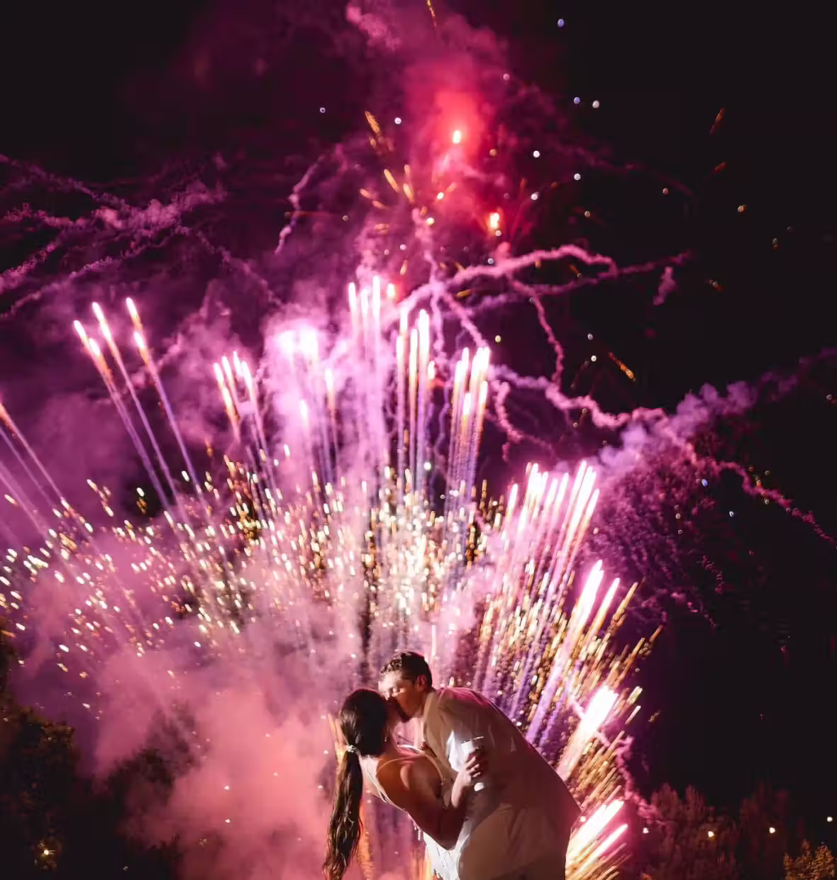 Couple kissing in front of vibrant pink and purple fireworks at night.