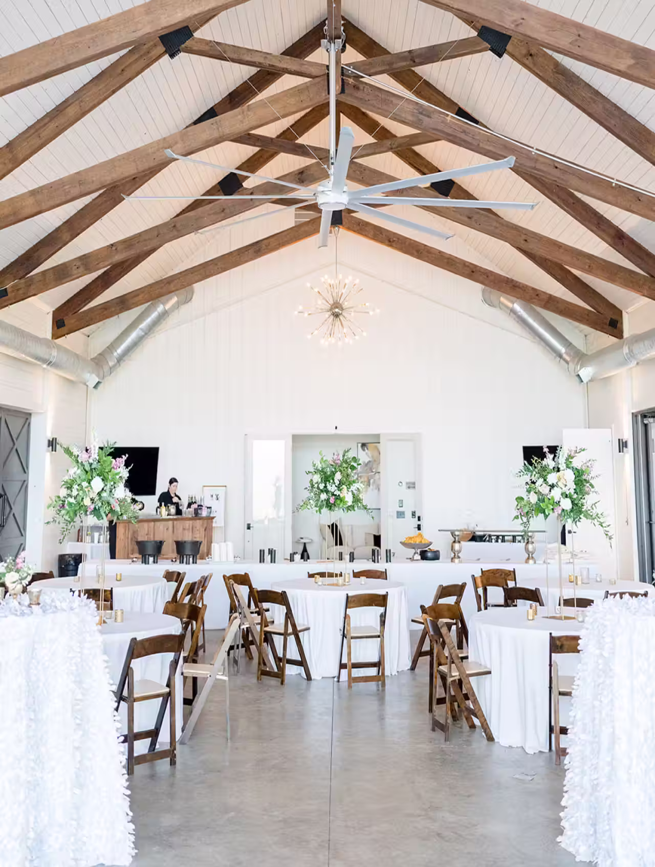 Bright event hall with wooden chairs around round tables covered in white linens and floral centerpieces under exposed wooden beams and a large ceiling fan.