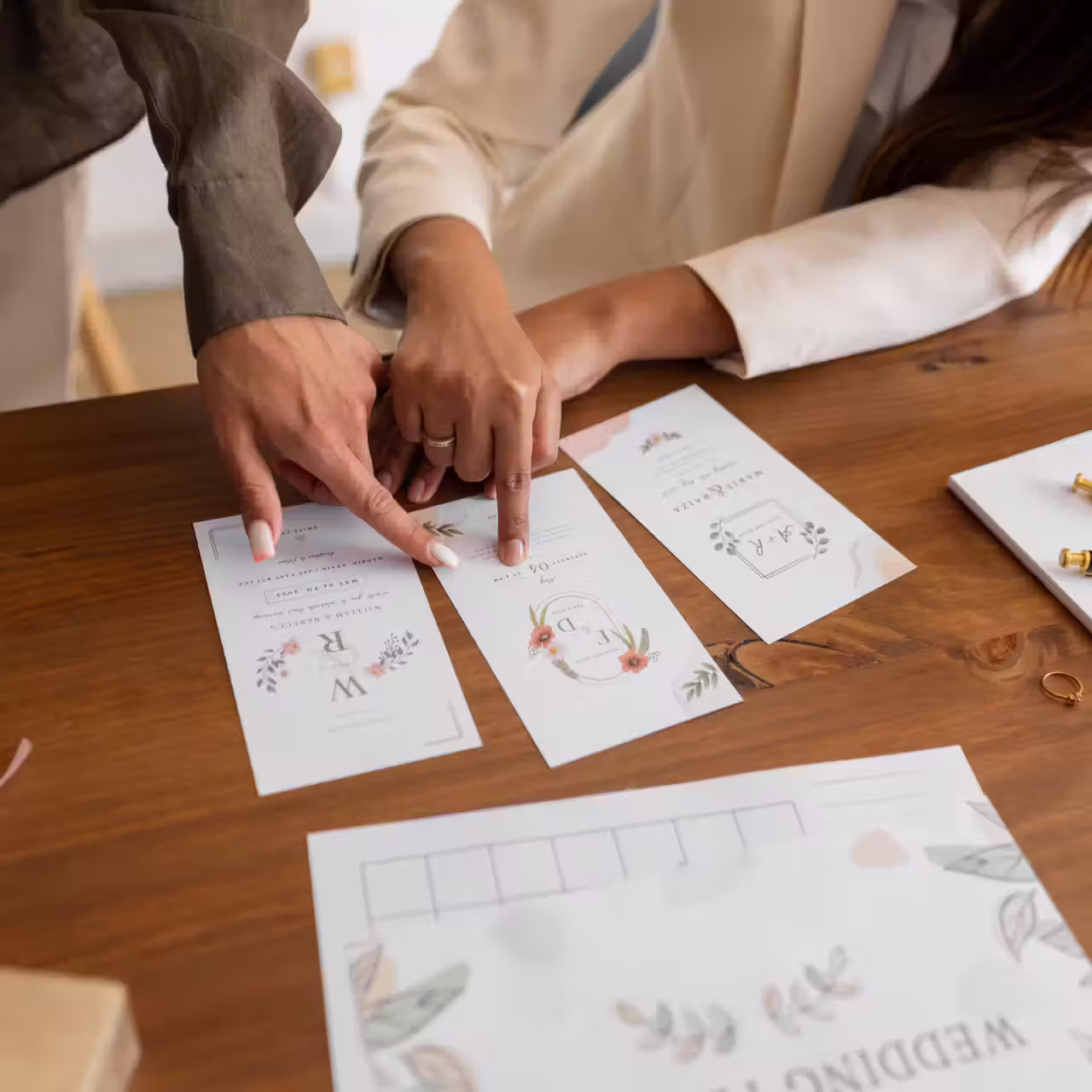 Two people pointing at floral-themed wedding invitation cards arranged on a wooden table.