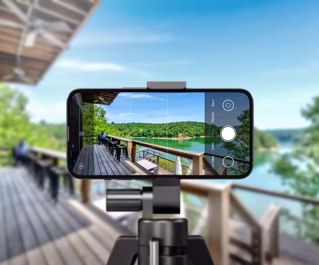 Smartphone on a tripod capturing a scenic view of a wooden deck overlooking a lake and forested hills under a clear blue sky.