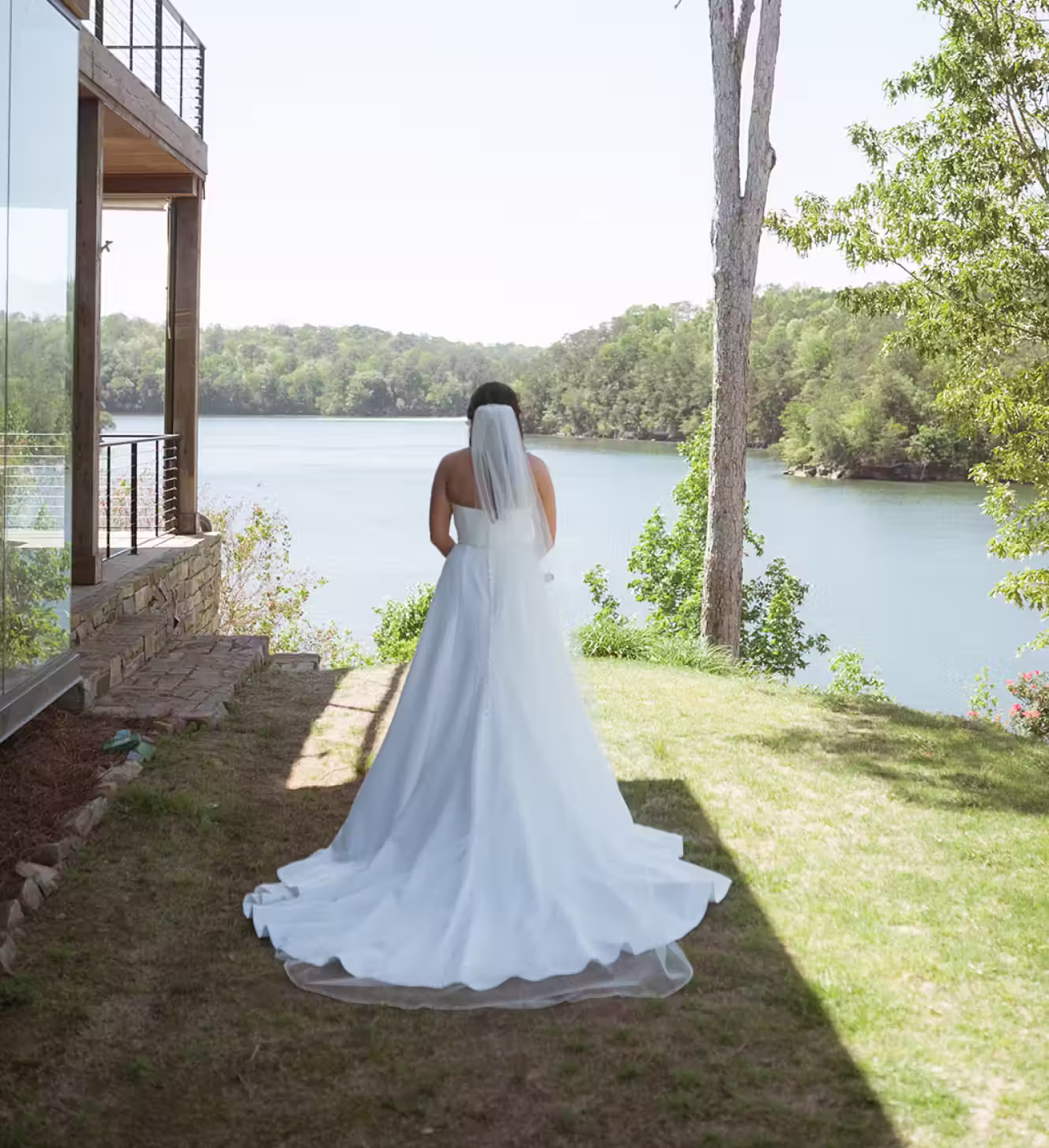 Bride in a white dress and veil standing on grass facing a lake surrounded by trees.