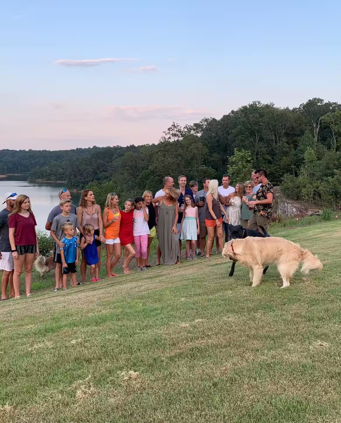 Large family group standing on grassy hill near a lake with trees in the background, two dogs nearby.