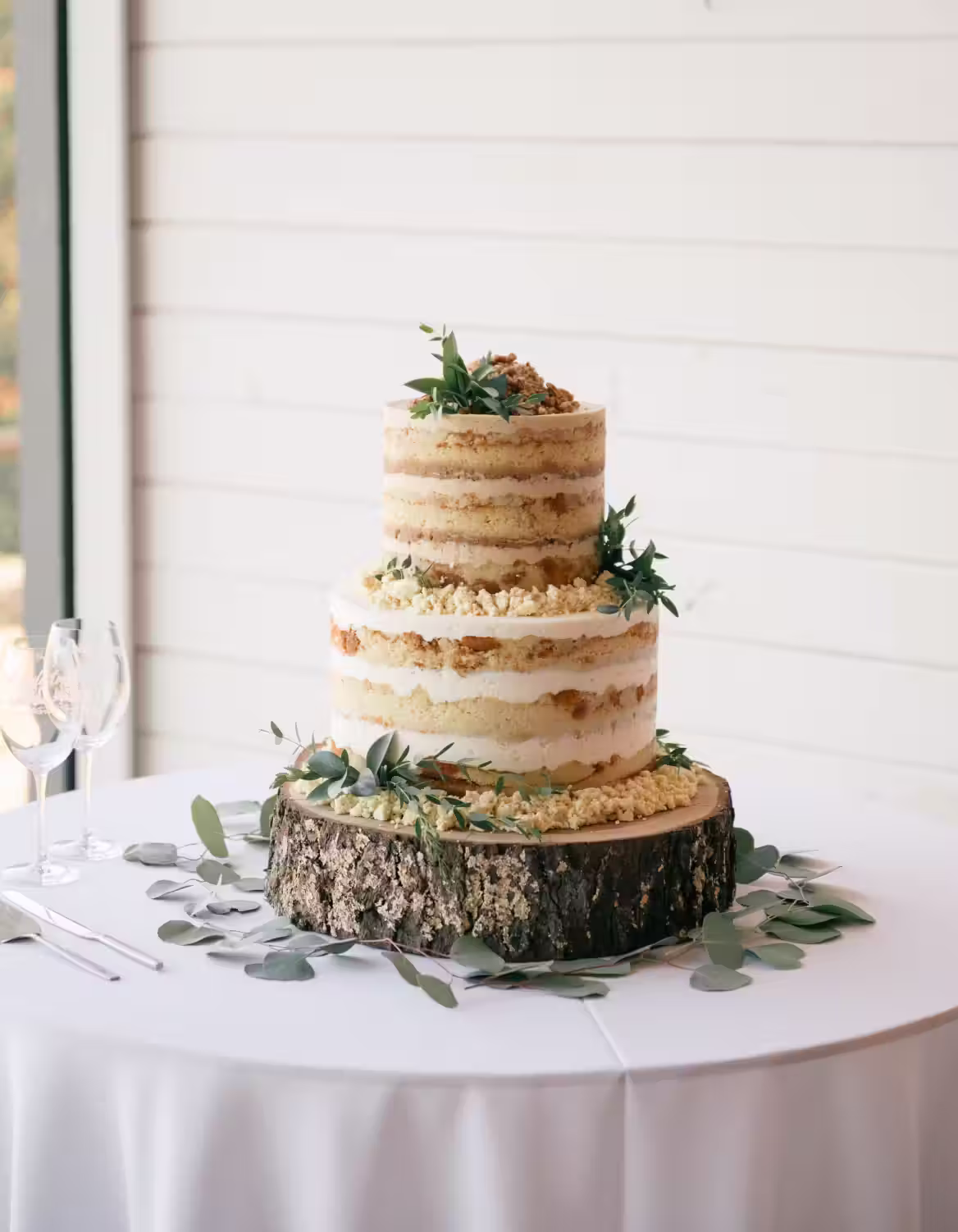 Two-tier naked cake decorated with greenery and crumbs, displayed on a wooden slab with eucalyptus leaves on a white tablecloth.