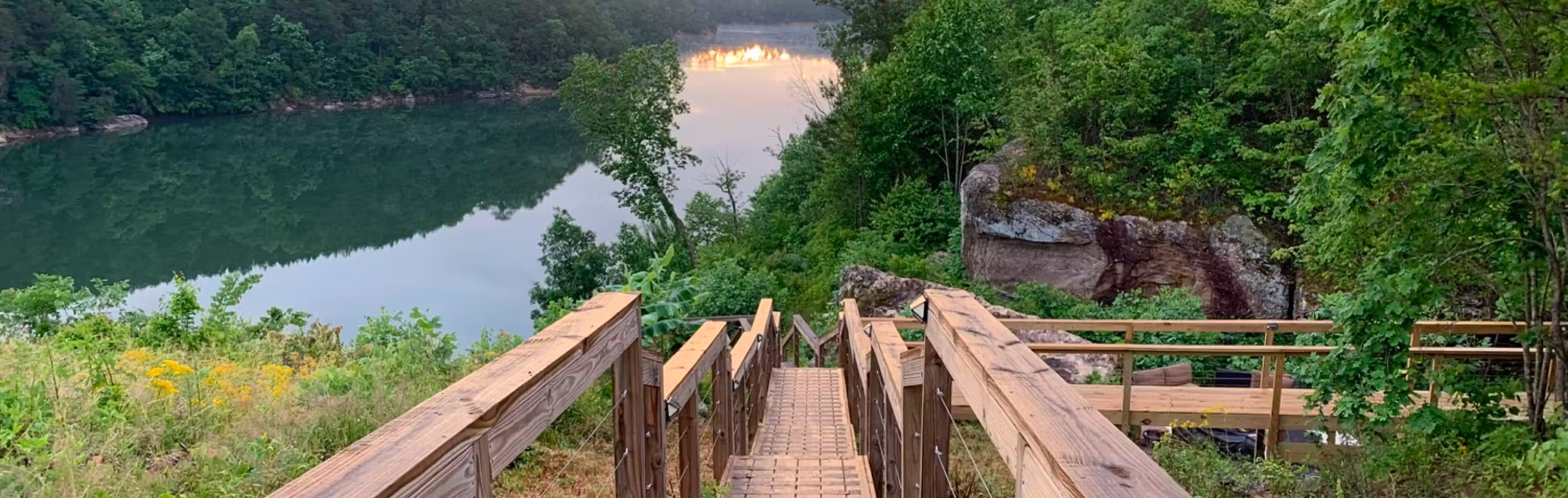 Wooden staircase leading down to a peaceful lake surrounded by dense green trees and rocky cliffs at sunset.