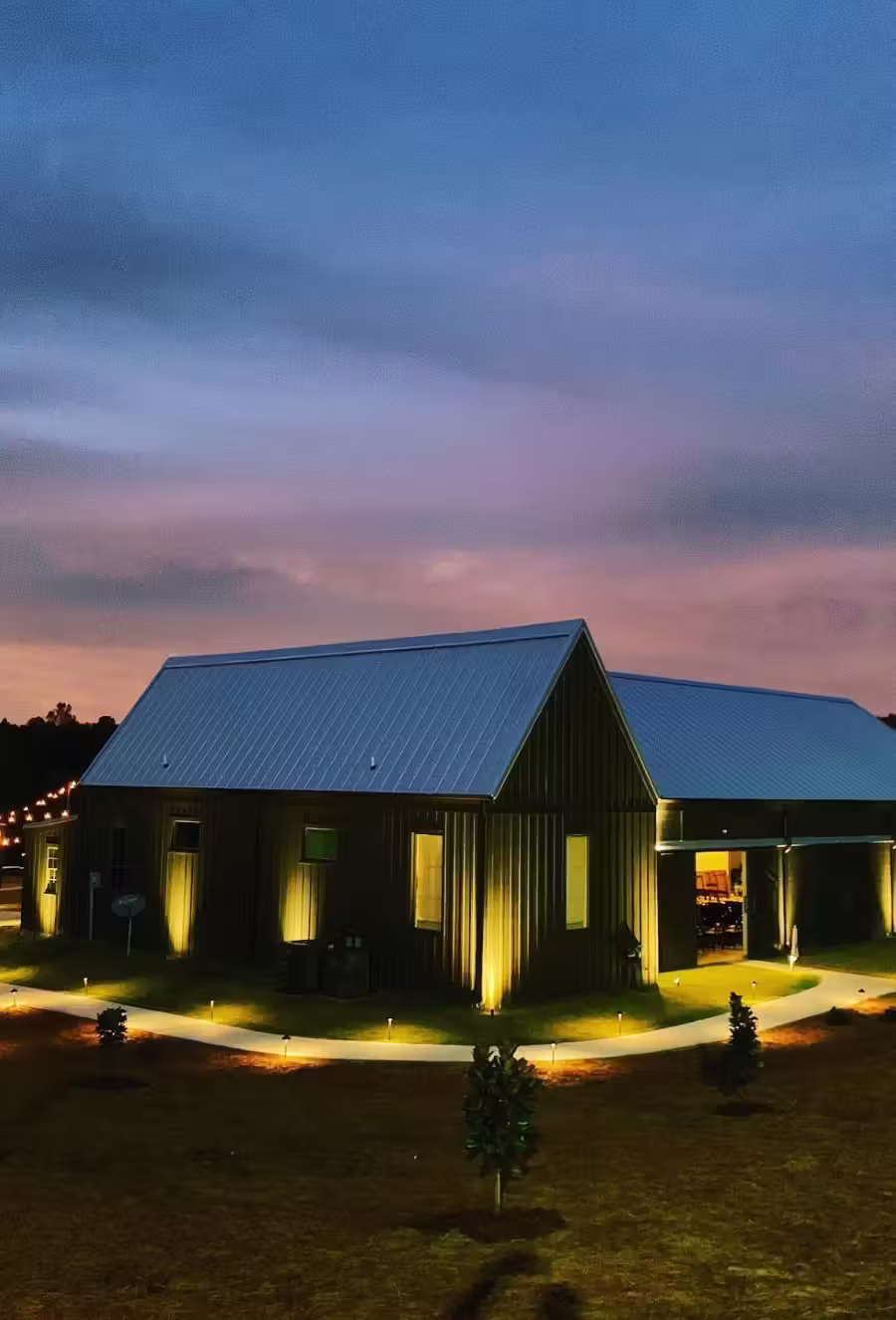 Barn with metal roof illuminated by warm lights along a curved walkway at dusk with a colorful sky.