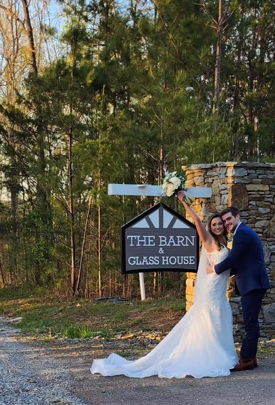 Bride and groom hugging and smiling in front of a stone structure with a sign reading 'The Barn & Glass House' surrounded by trees.