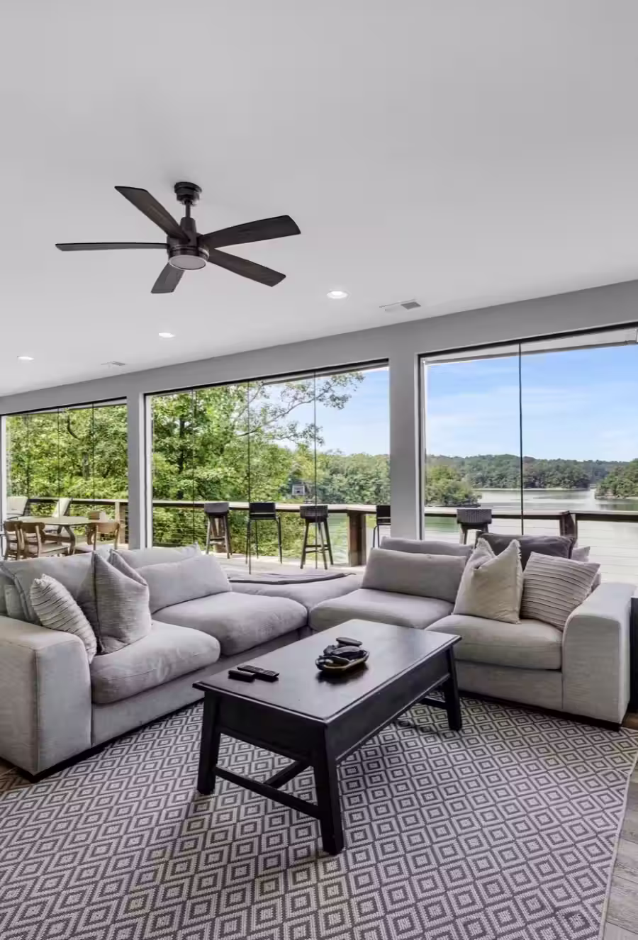 Modern living room with gray sectional sofa, black coffee table, and large windows overlooking green trees and a river.