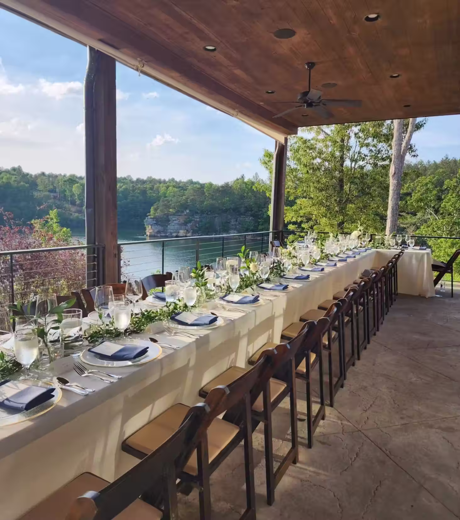 Long outdoor dining table set with glassware, plates, and greenery overlooking a lake and forest.