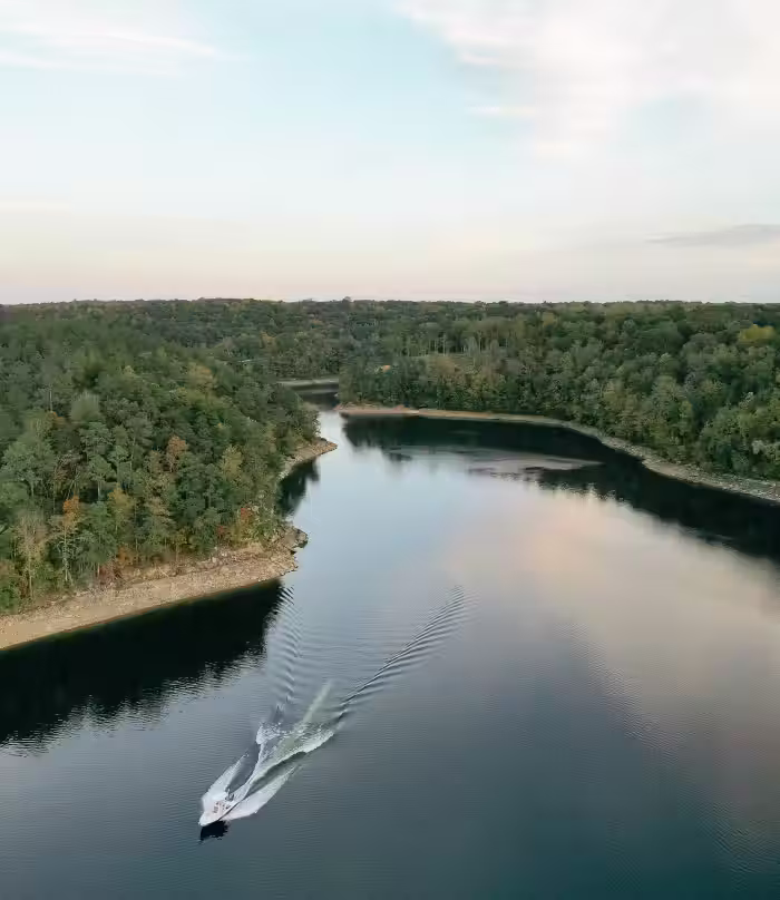 A small motorboat creating waves as it moves along a river surrounded by dense forest.