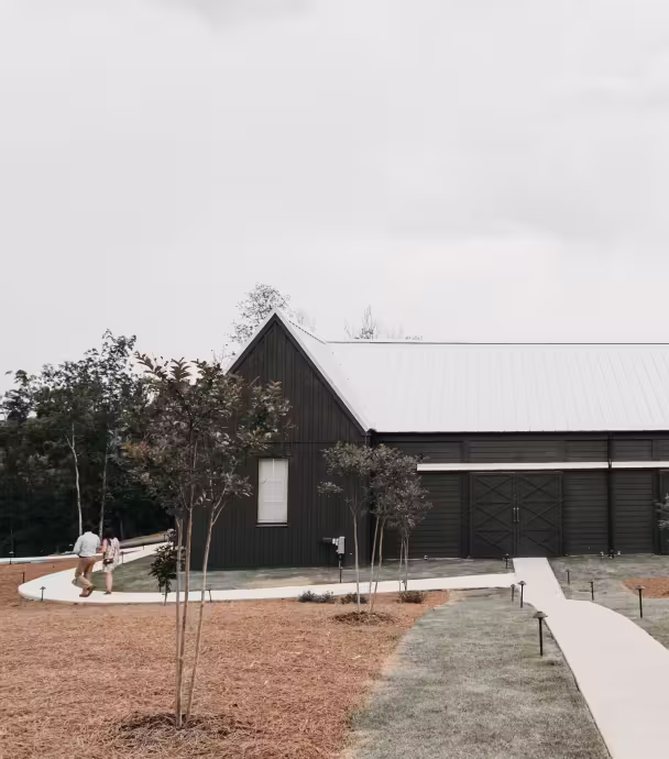Modern black barn-style building with white roof, small trees in front, and two people walking on the curved concrete pathway.