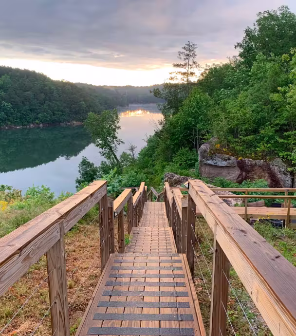 Wooden stairs leading down to a calm river surrounded by green trees under a partly cloudy sky at sunset.