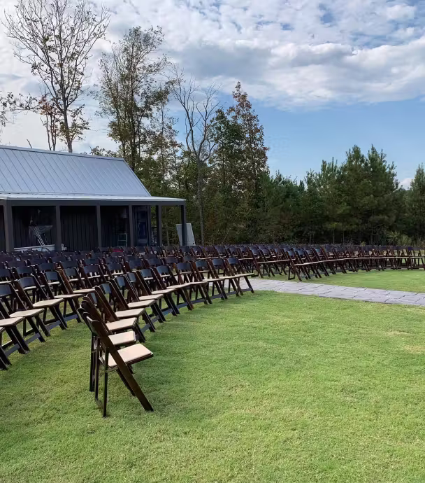 Rows of wooden folding chairs arranged on green grass outdoors in front of a pavilion with trees and a cloudy sky in the background.
