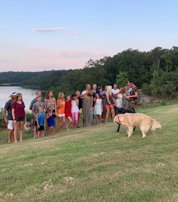 Group of people of various ages standing on a grassy hill near a lake with trees, watching two dogs playing.