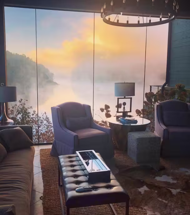 Cozy living room with armchairs, a leather ottoman, and lamps, featuring a large window showing a foggy lake at sunrise.