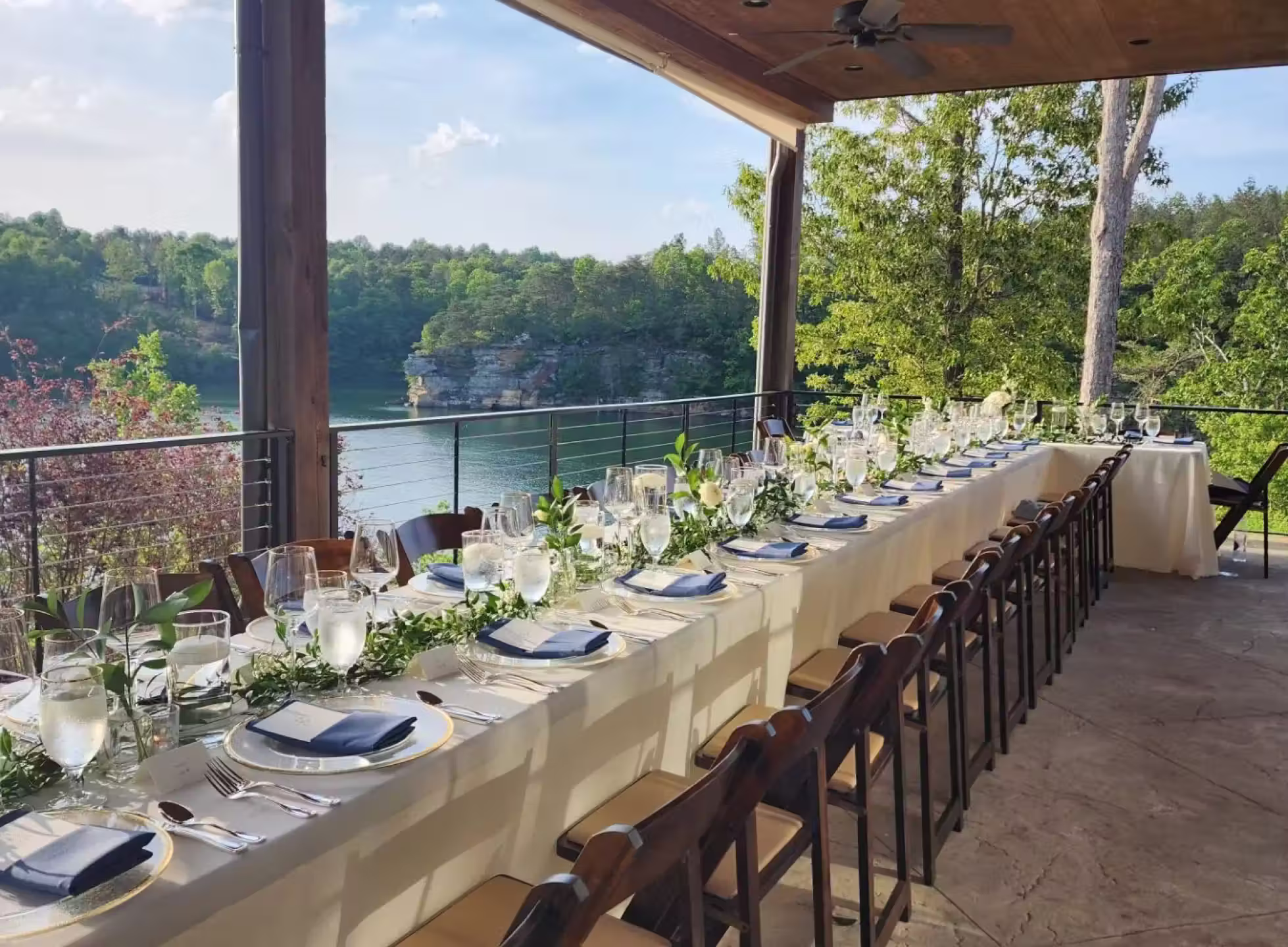 Long outdoor dining table set with plates, glasses, silverware, and greenery centerpiece on a covered patio overlooking a lake and forest.