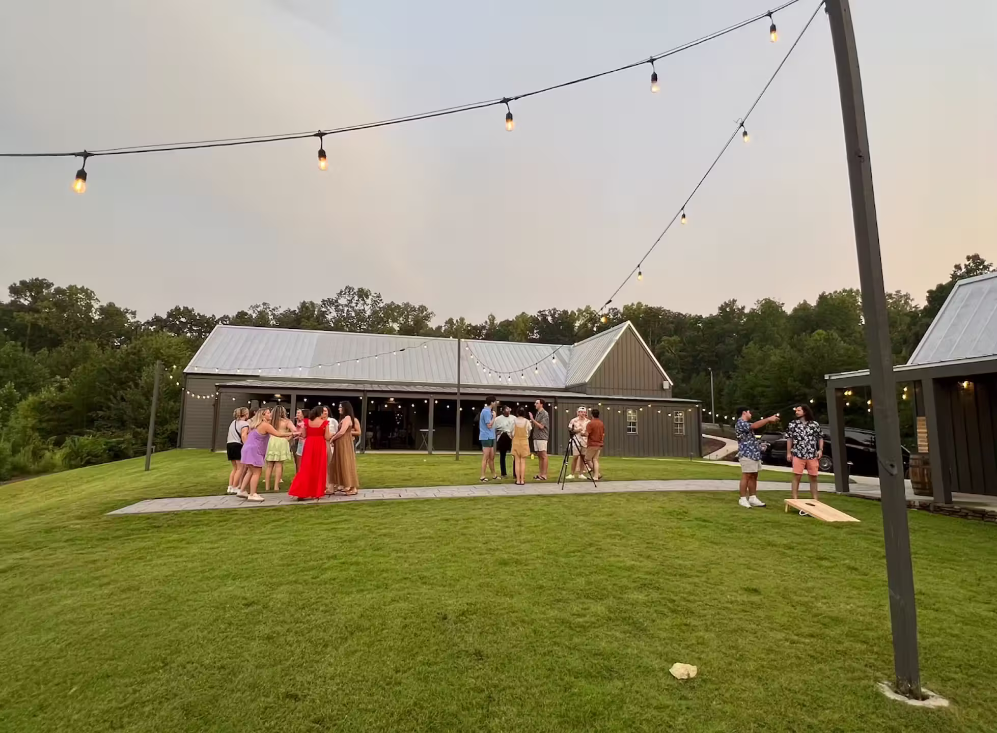 Group of people socializing on a manicured lawn outside a large barn-style building with string lights overhead at dusk.