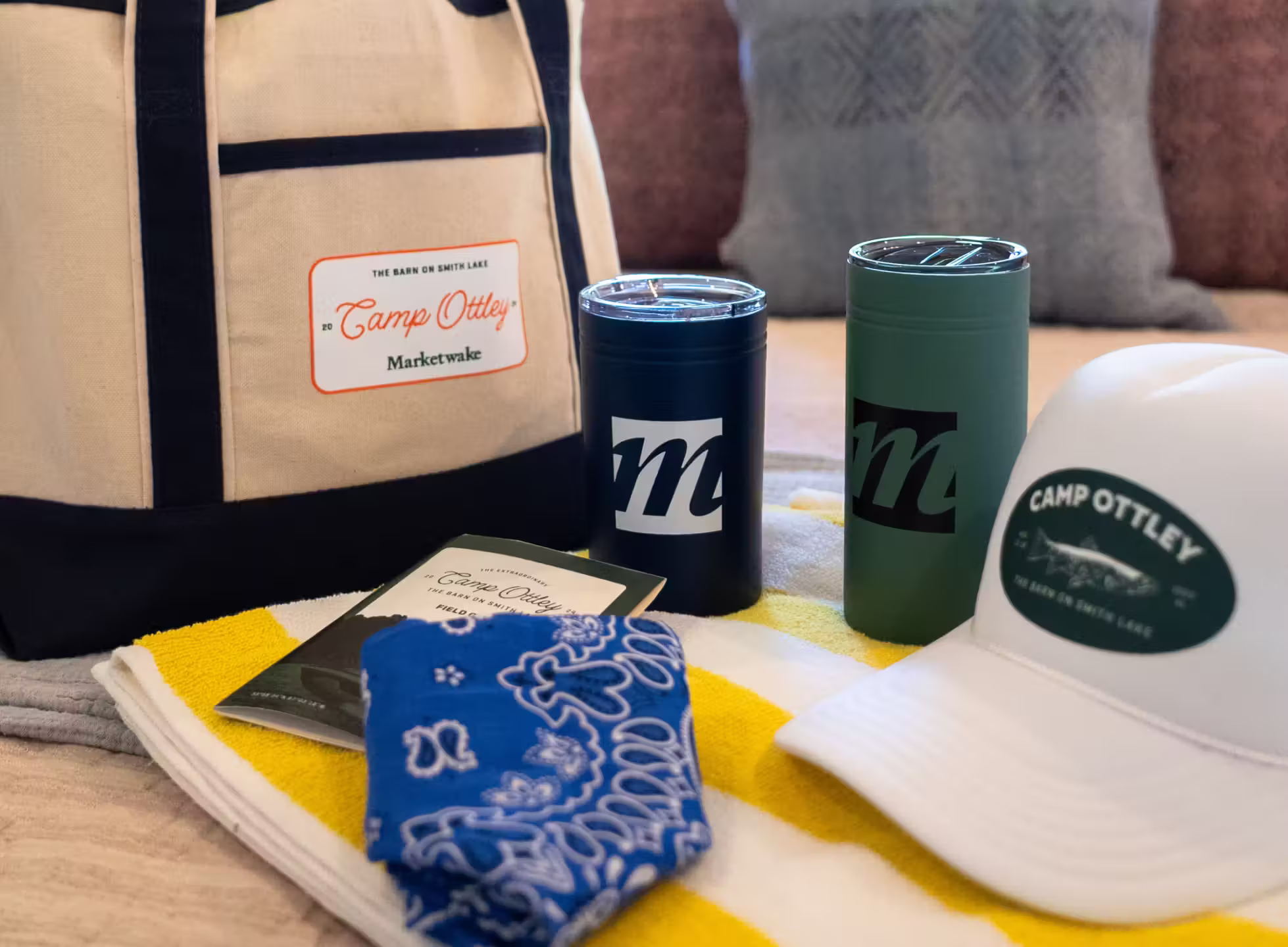 Collection of Camp Ottley merchandise including a tote bag, two insulated cups, a white hat, a blue bandana, and a yellow and white towel.