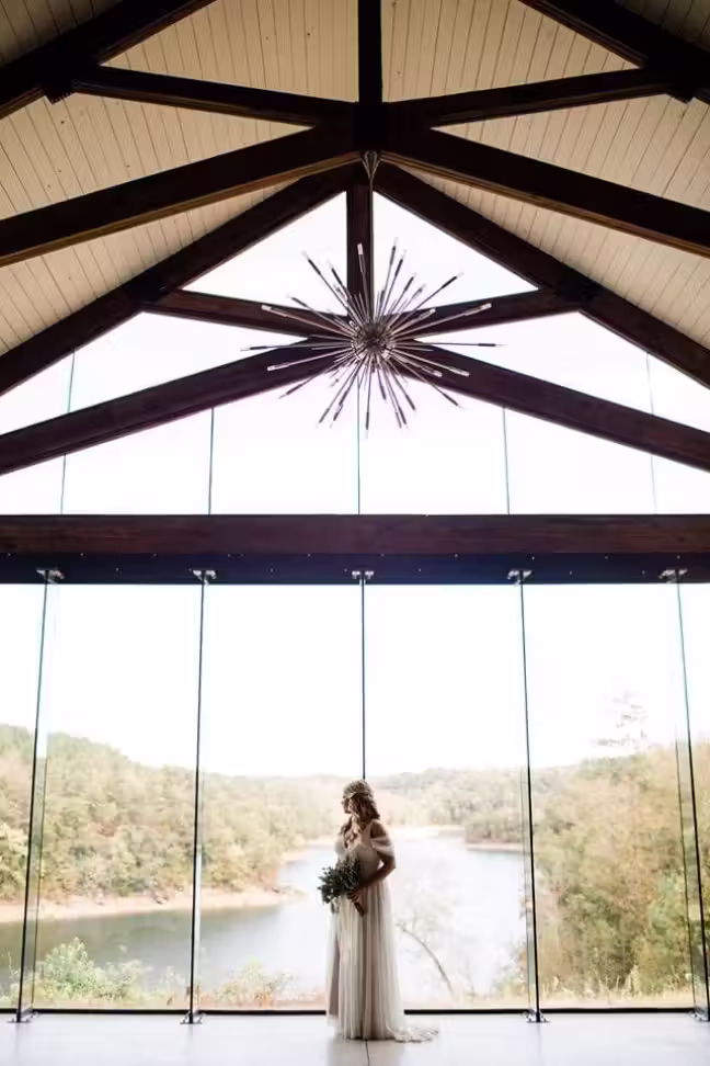 Bride in a white dress holding a bouquet, standing in front of large floor-to-ceiling windows overlooking a river and forest landscape, inside a venue with a wooden beam ceiling and modern chandelier.
