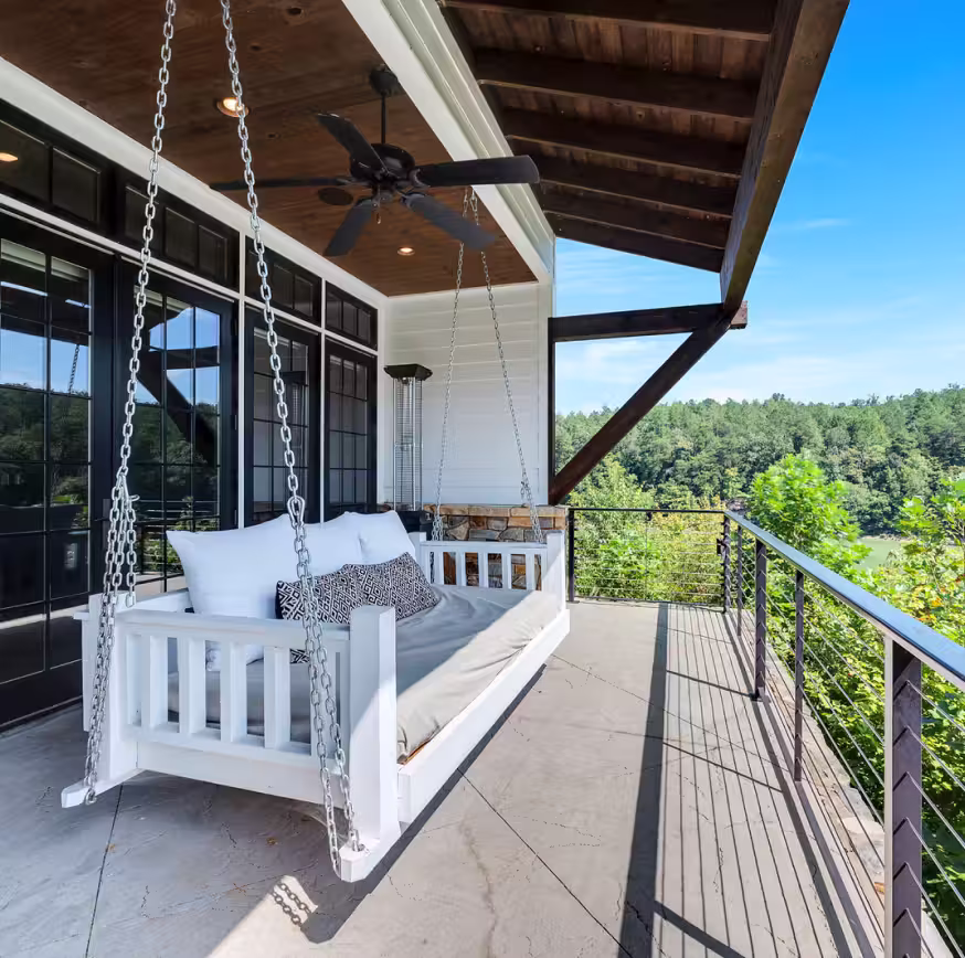 White wooden porch swing with cushions hanging on chains under a wooden roof on a balcony overlooking green trees and a clear blue sky.