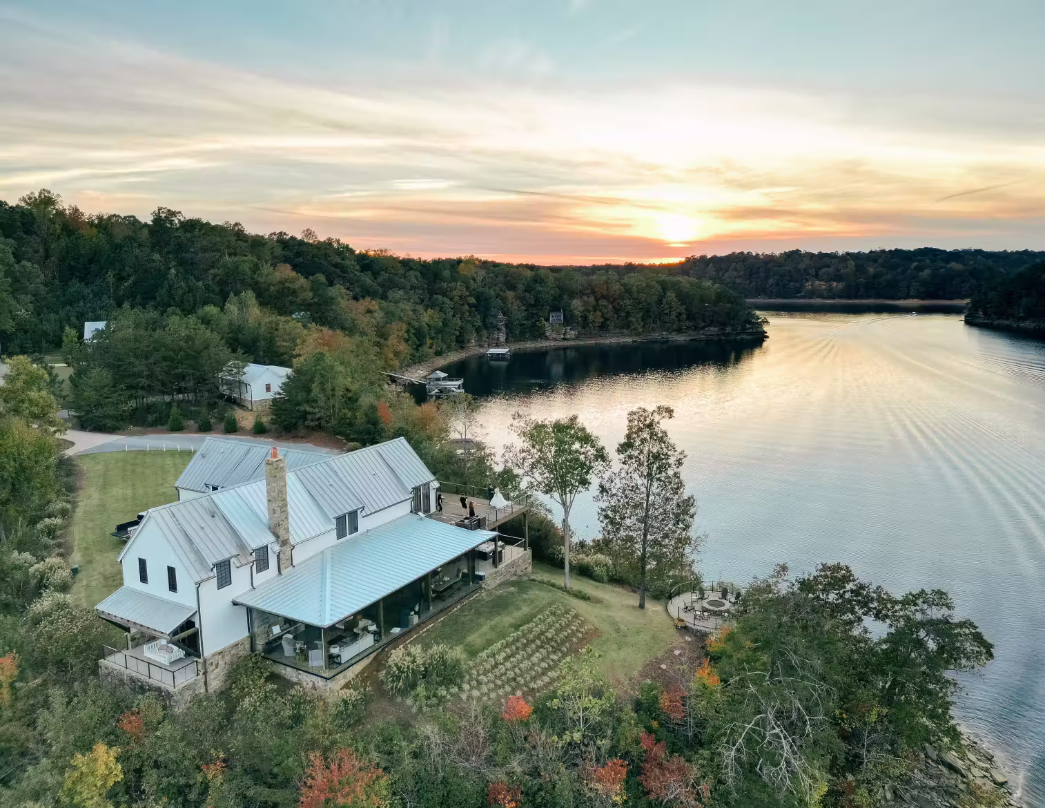 A large lakeside house surrounded by trees with a view of a calm lake at sunset.