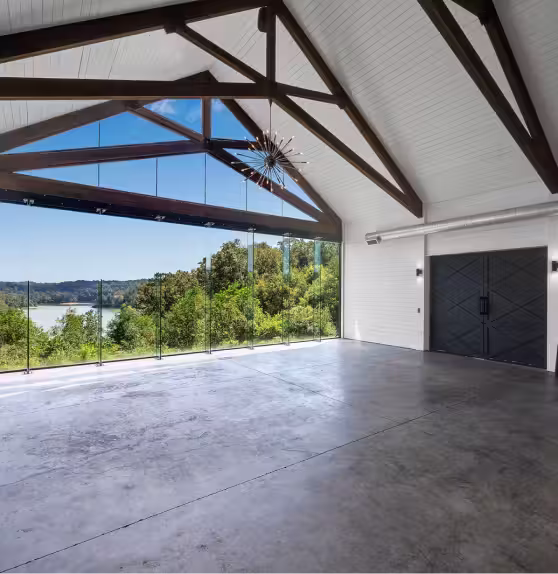 Spacious modern hall with large glass windows overlooking a lake and surrounding green trees, featuring exposed dark wooden beams and concrete floor.