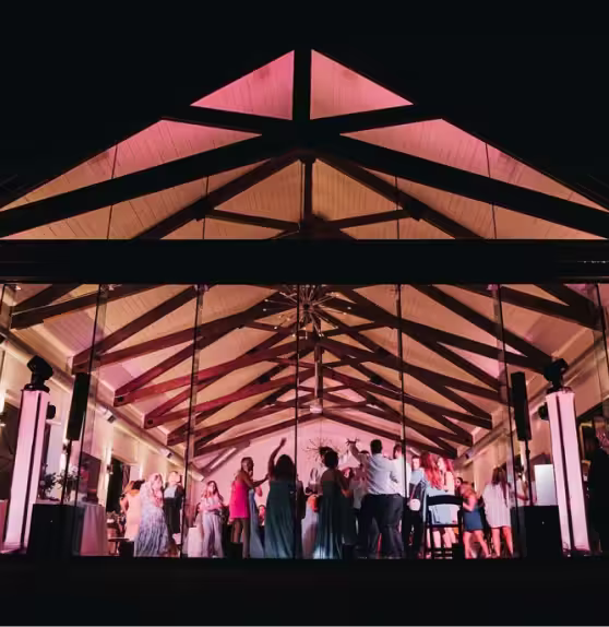 People dancing and celebrating inside a warmly lit venue with a high triangular wooden ceiling at night.