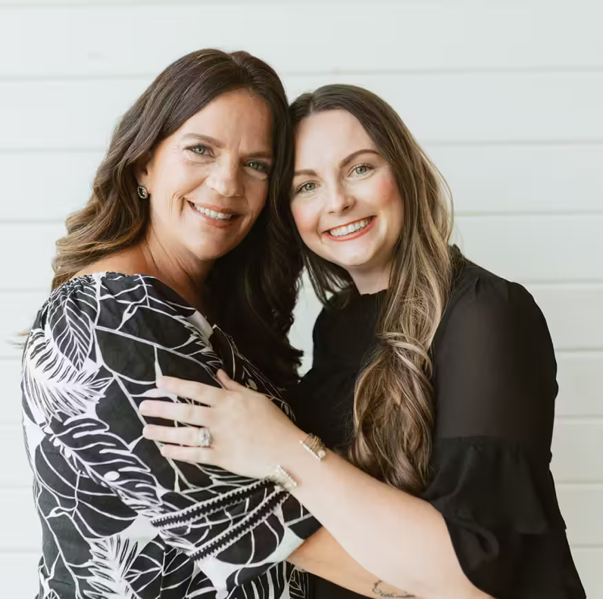 Two smiling women embracing each other, one in a black and white patterned top and the other in a black blouse, standing against a light-colored wall.