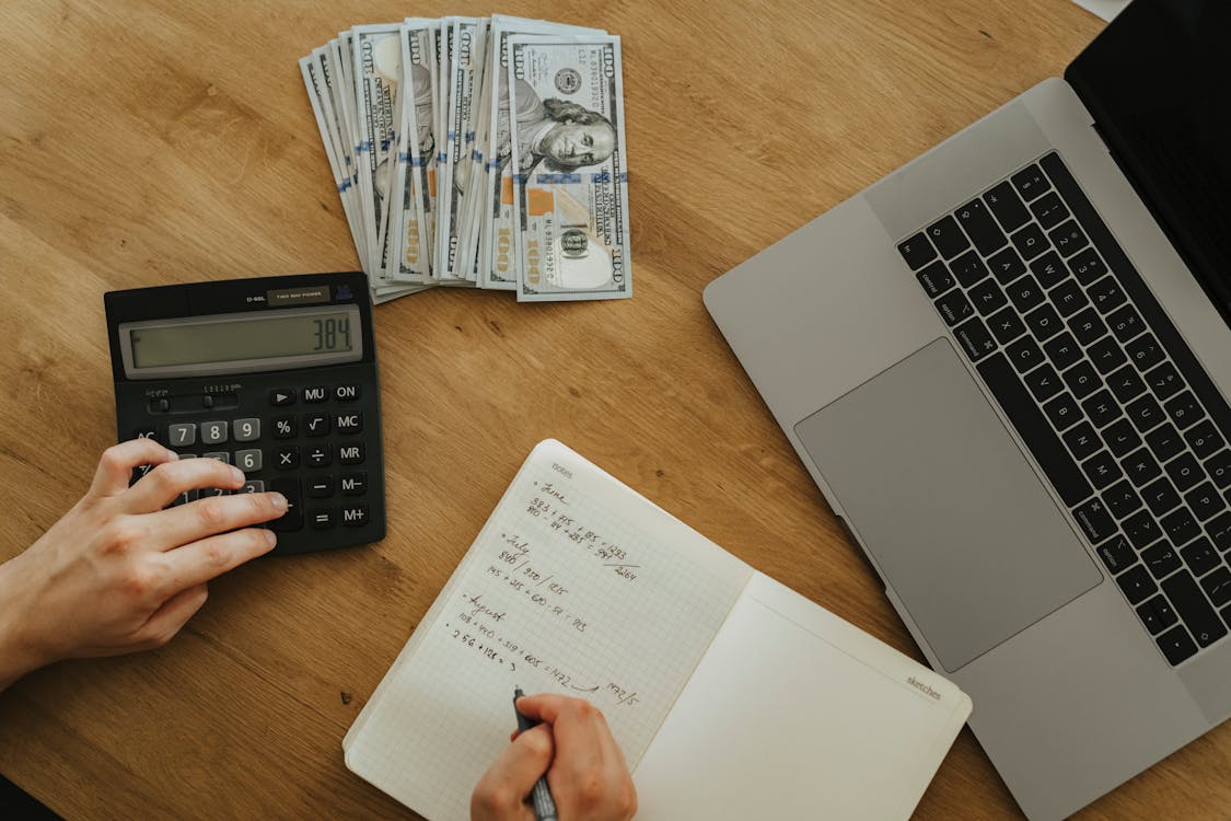 Person counting cash with a calculator, notebook and laptop on a wooden desk, representing the manual reconciliation many small dojo owners still do at month-end