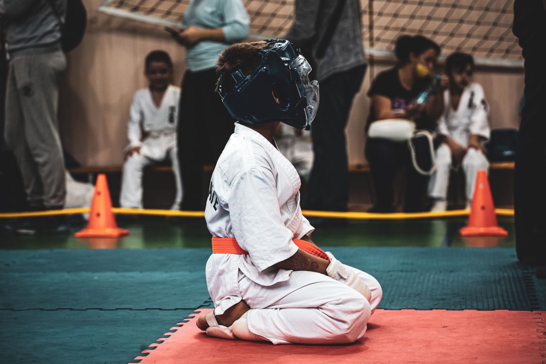 A young martial arts student in a gi and headgear kneeling on the mats at a dojo event, with classmates and parents in the background