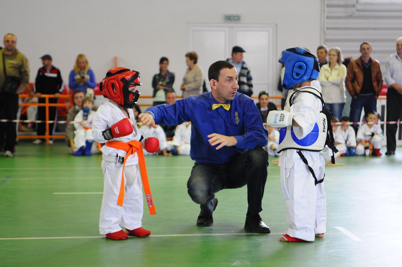 Referee starting a children's karate match while parents and young competitors watch from the side of the mat