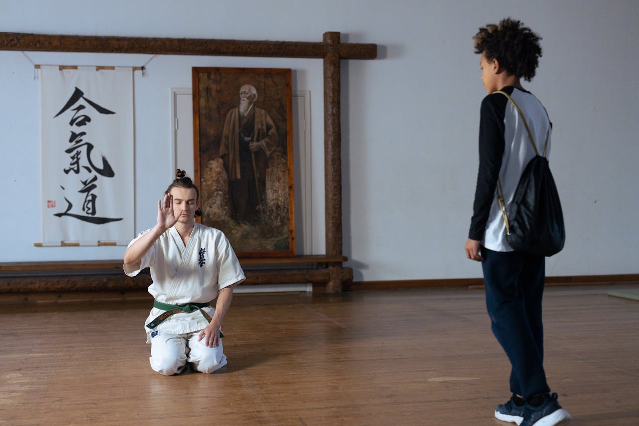 A martial artist kneeling in a white gi greets a boy arriving at an Aikido dojo, with Aikido calligraphy and a portrait of the founder on the wall