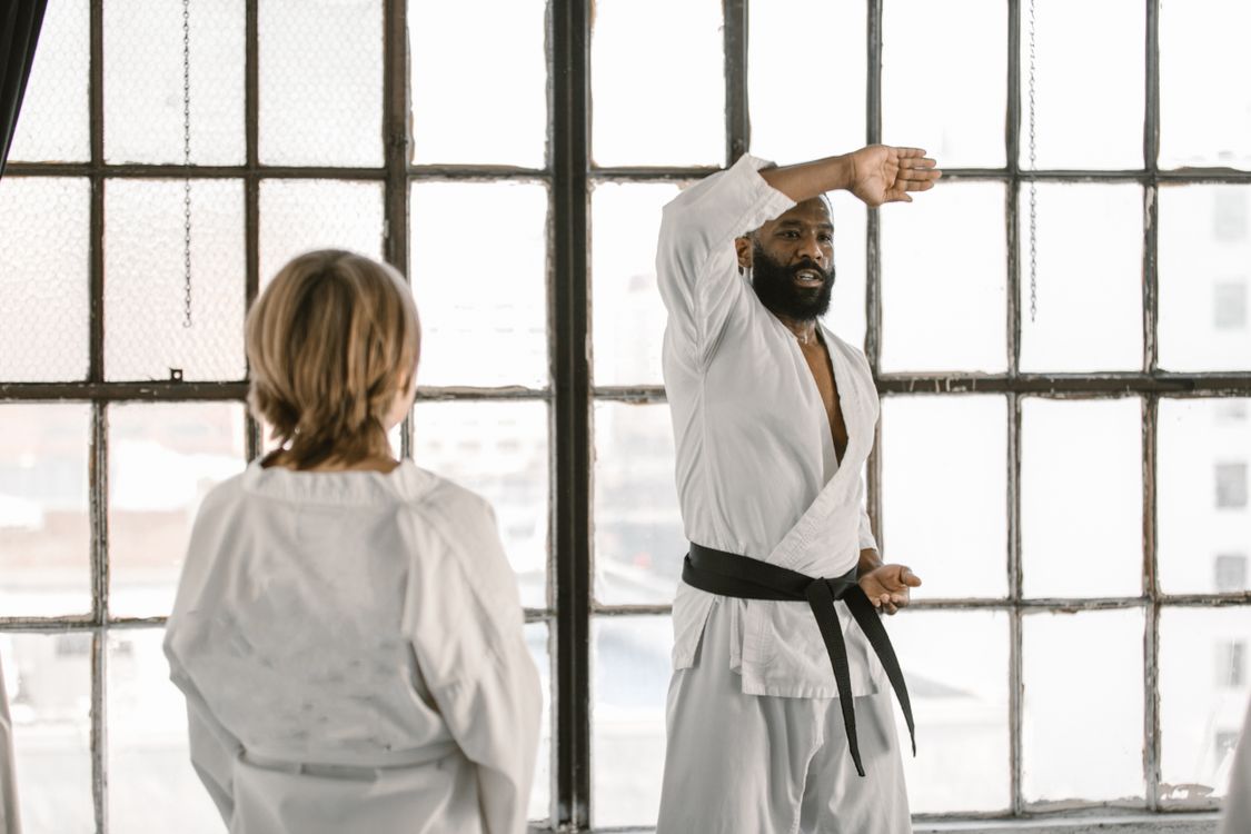 A black belt instructor demonstrating a technique to a student in a bright dojo with large windows