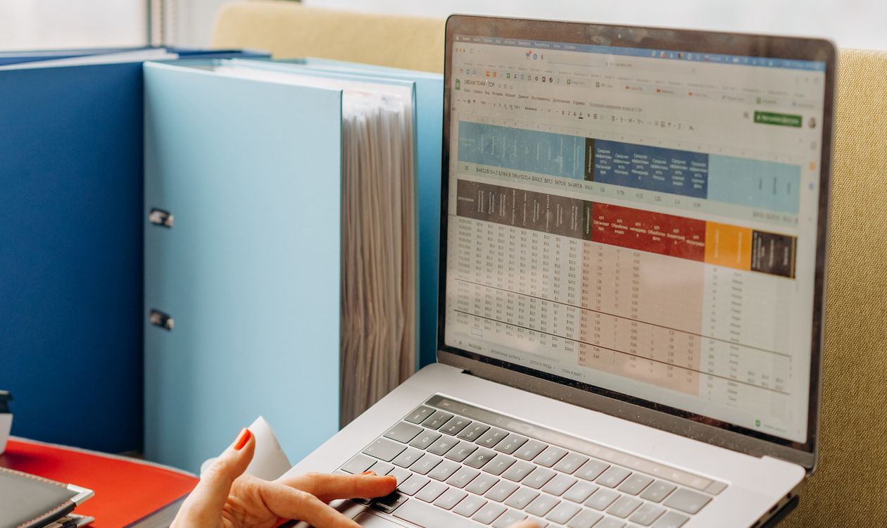 Someone working at a laptop showing a dense colour-coded spreadsheet, with ring binders of paper files stacked beside the desk