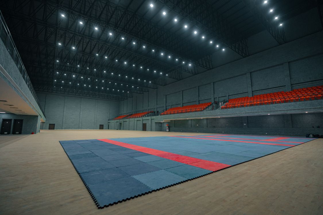 An empty sports hall with blue and red tatami laid out and rows of empty spectator seating, hours before a martial arts event begins