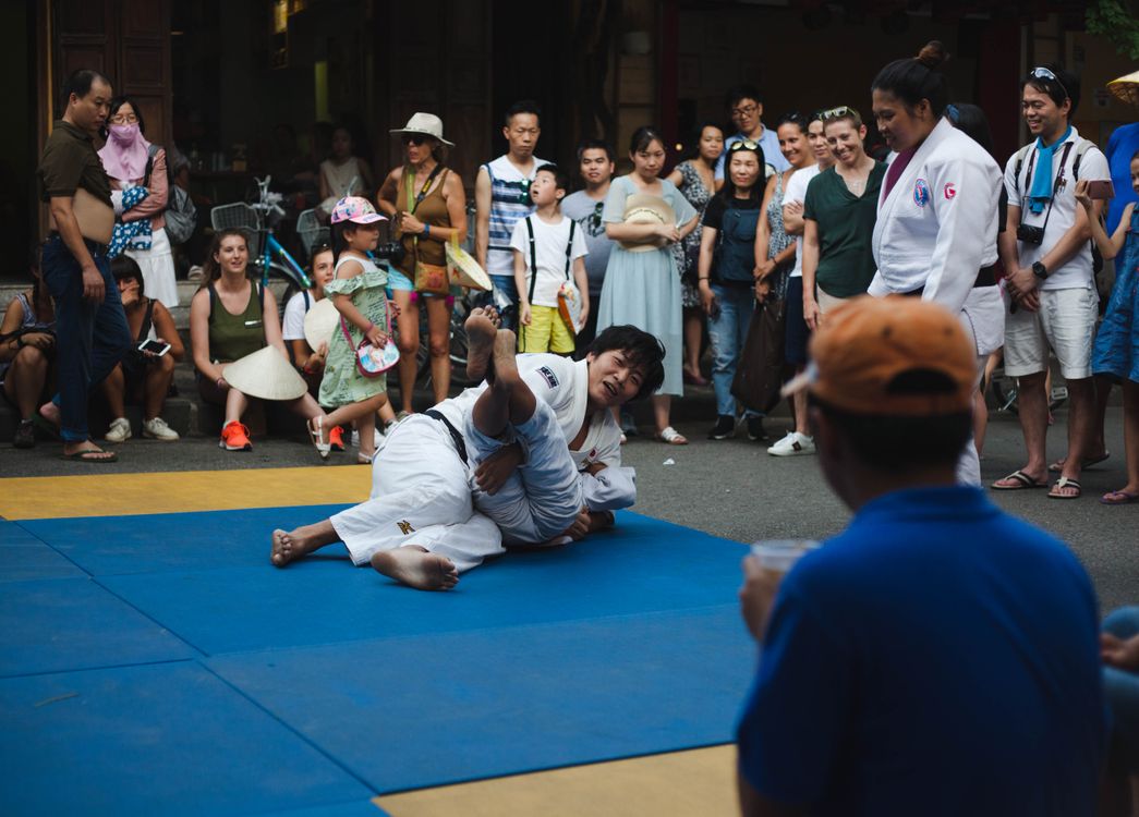 Two judoka grappling on blue and yellow mats laid in a street, surrounded on three sides by a standing crowd of onlookers at a public demonstration