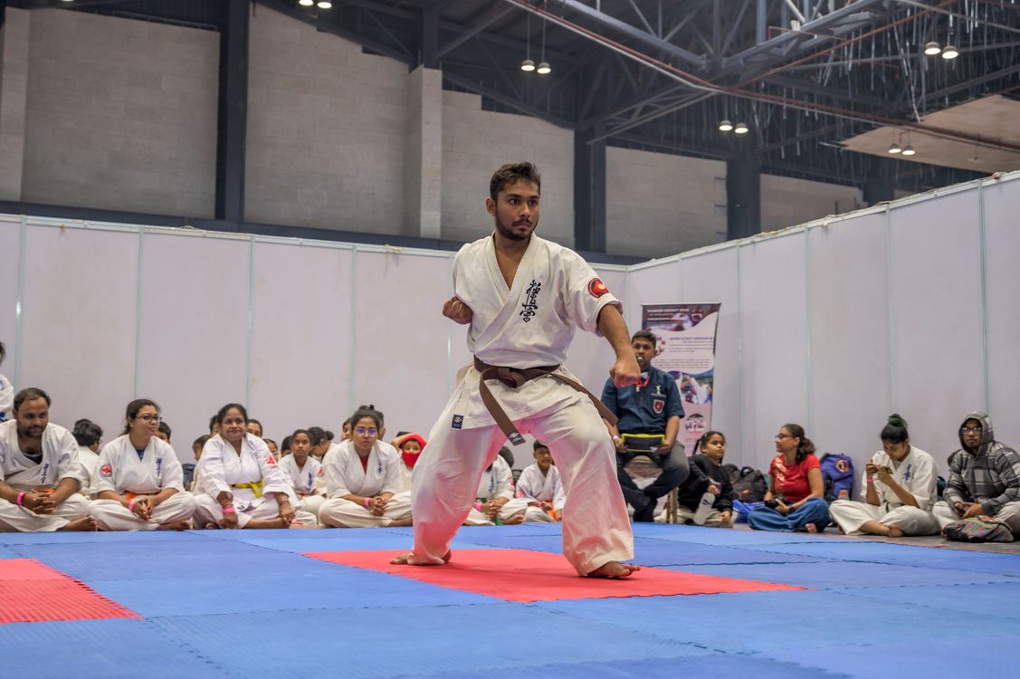 A karateka demonstrating a form on blue and red mats in a large sports hall while rows of students in white gi sit along the edge and watch, at a martial arts event in Kolkata, India