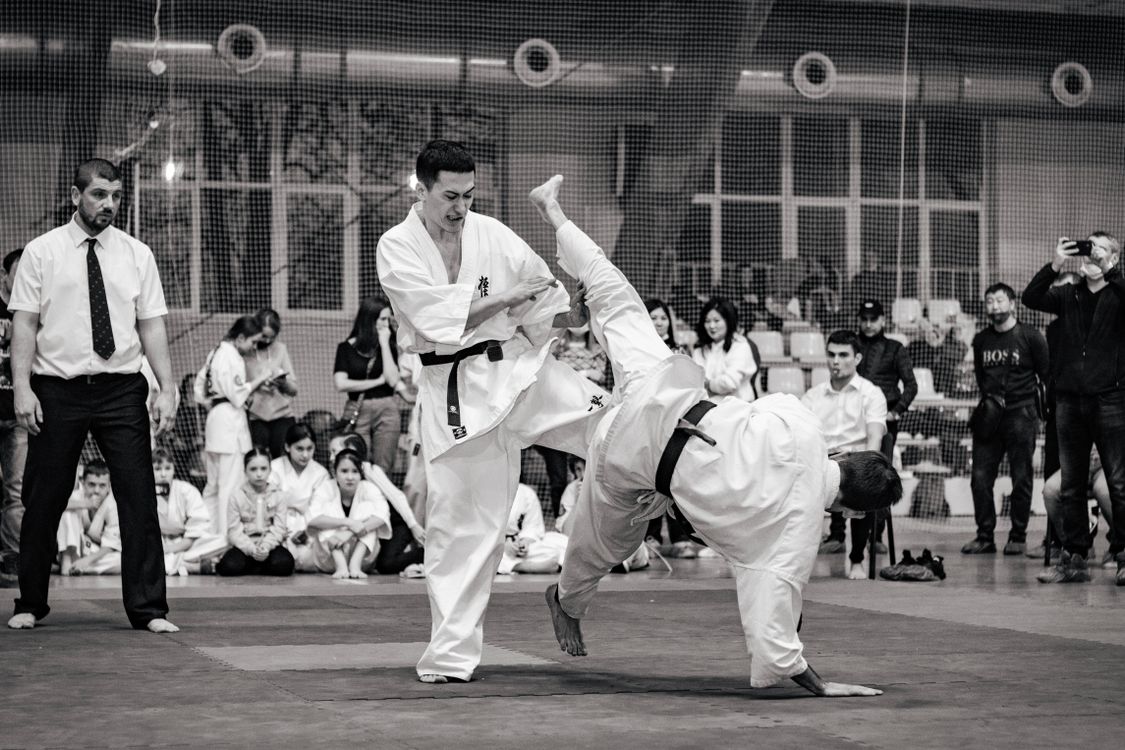 A karate bout at a tournament, with a referee in shirt and tie and rows of competitors and spectators watching from the edge of the mat, in black and white
