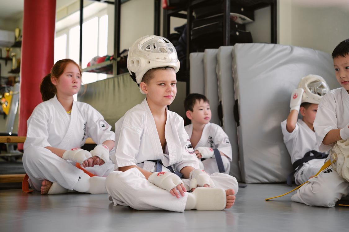 Several children in white karate gi wearing padded white headguards and hand mitts, sitting cross-legged on the floor waiting their turn at an event