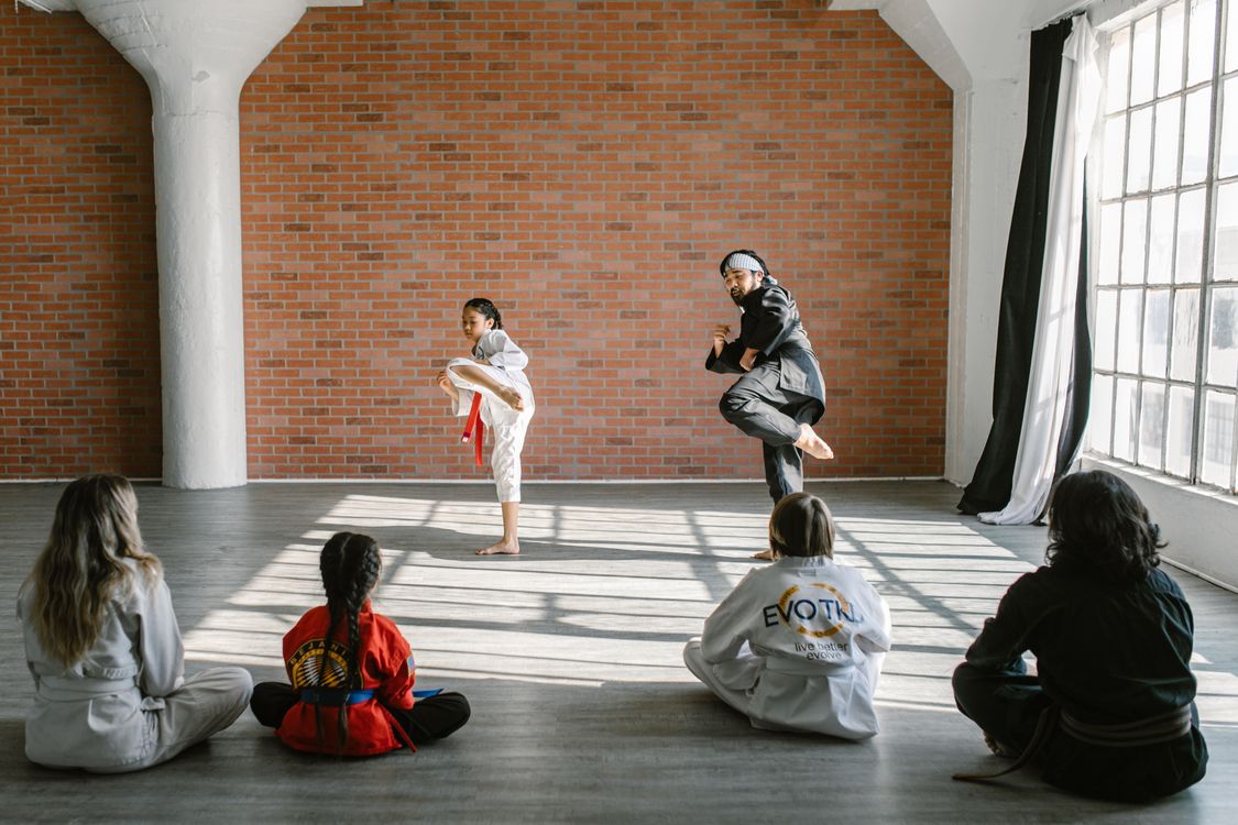 A martial arts instructor demonstrating a kick to a young student while the rest of the children's class watches from the mat