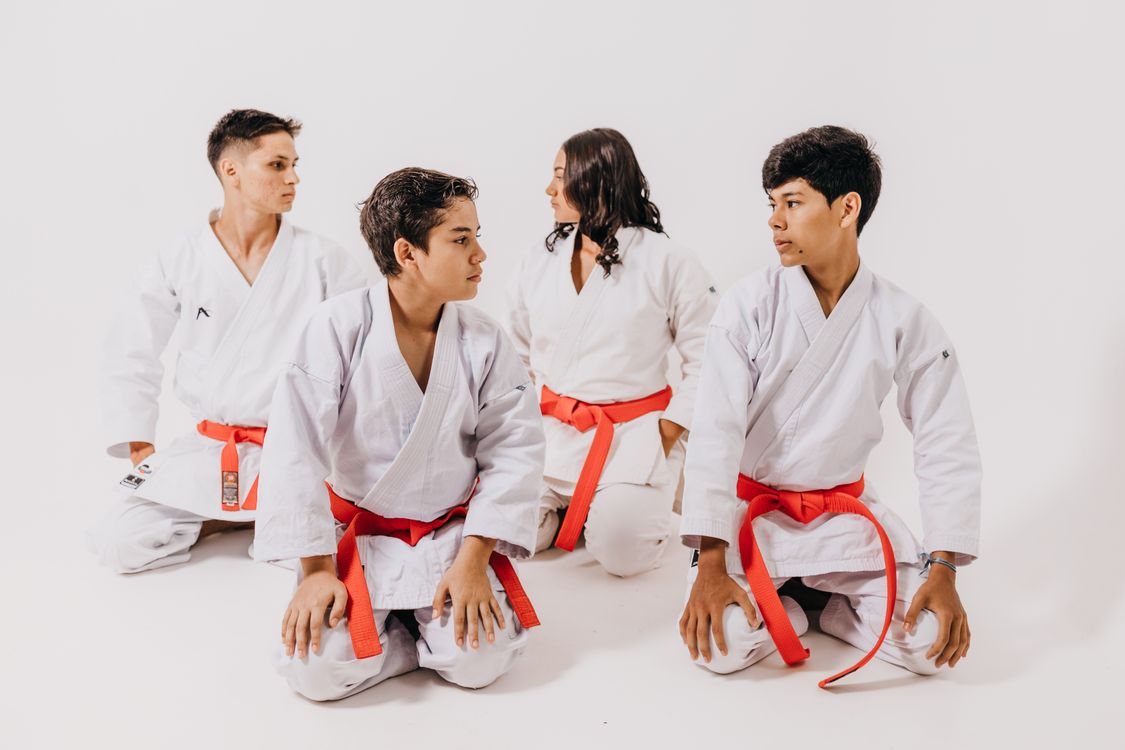 Four teenage karate students in white gi with red belts kneeling together on the mat