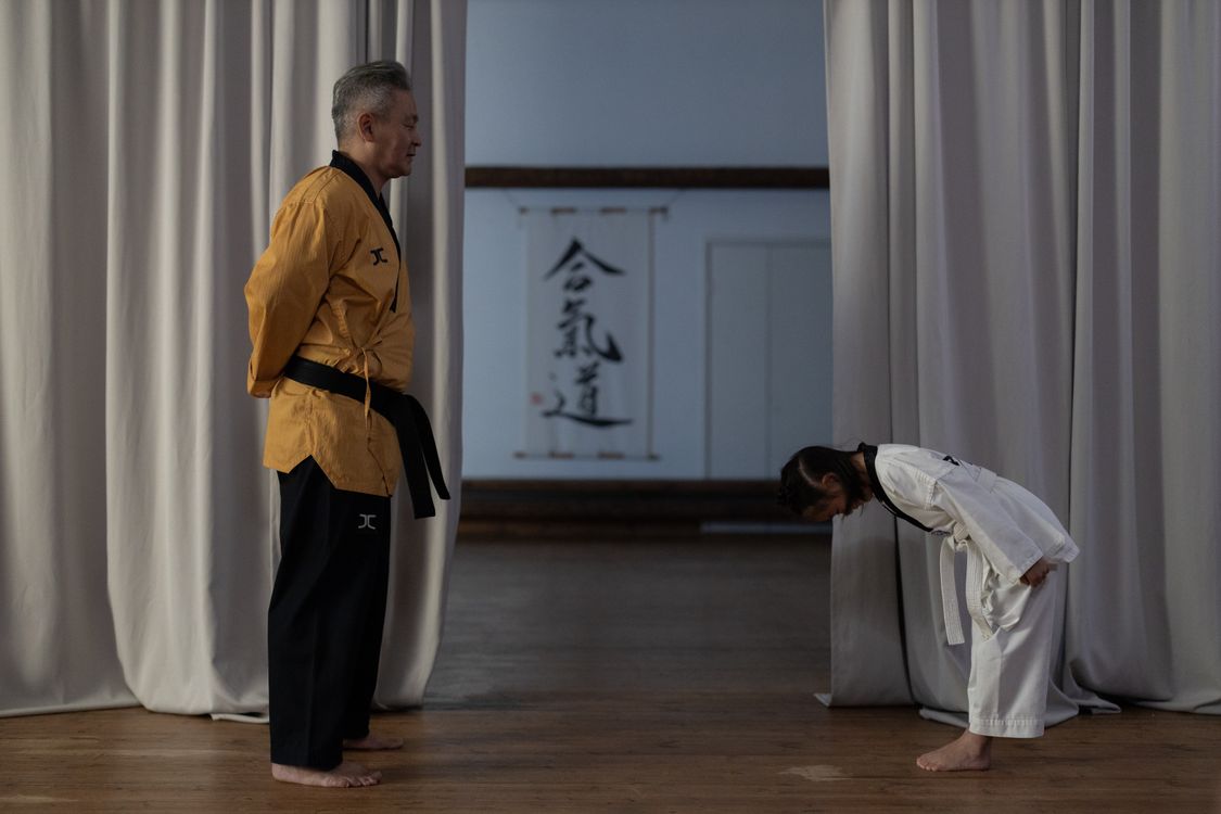 A young student in a white gi bows to her instructor at the start of class in a dojo hung with calligraphy