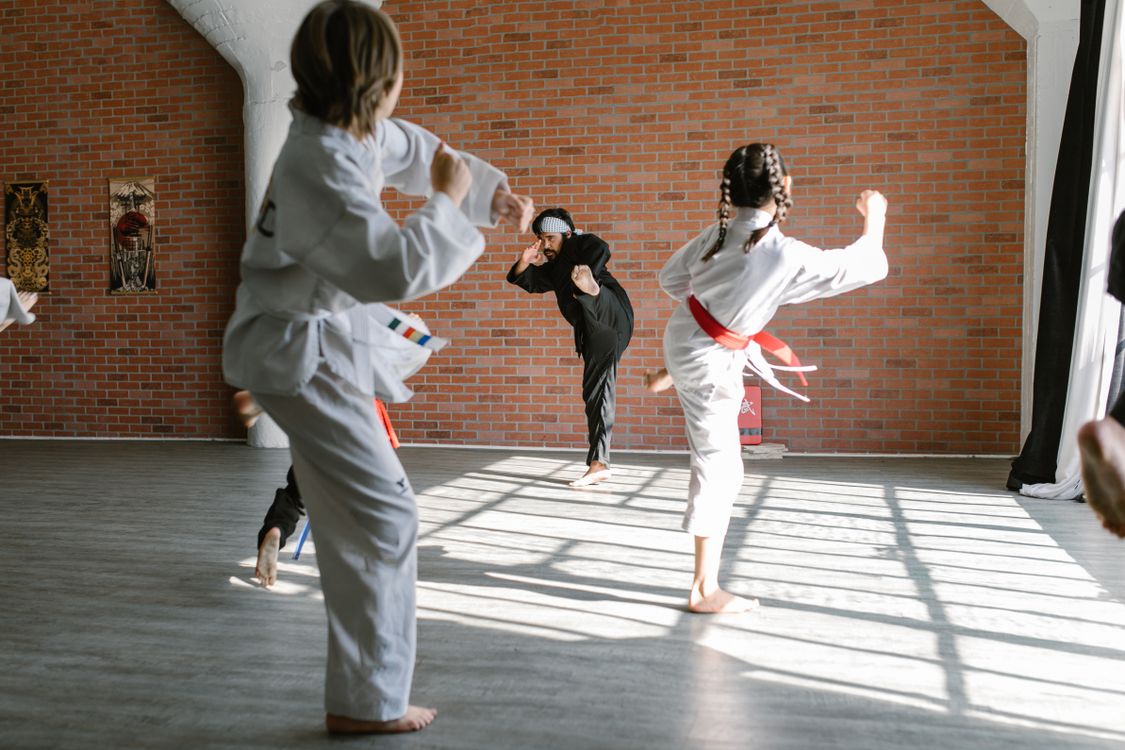 A group of children practise karate kicks in a bright dojo while their instructor leads from the front