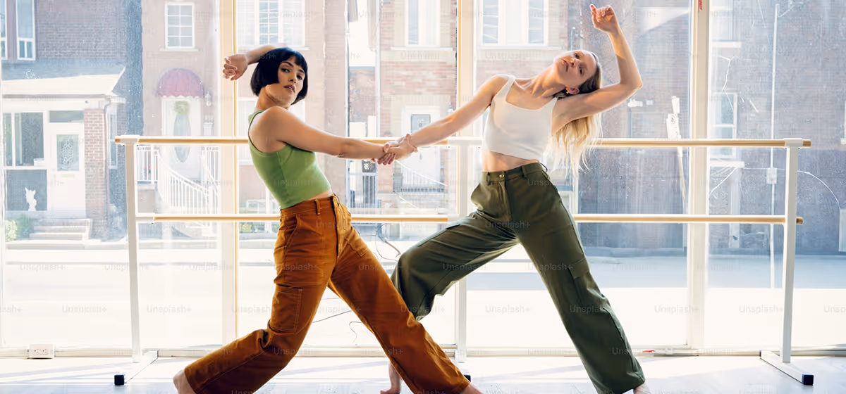 Twee vrouwen dansen samen in een lichte ruimte met grote ramen en houten dansbeugels.